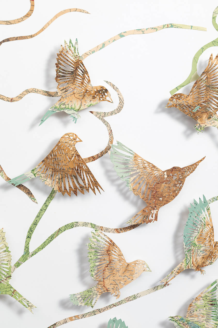 A close up detail of: Seeing The Future by Claire Brewster. A large paper sculpture of birds flying among winding, branch-like paths, hand-cut from pages of a vintage The Times Atlas in earthy green and brown tones.