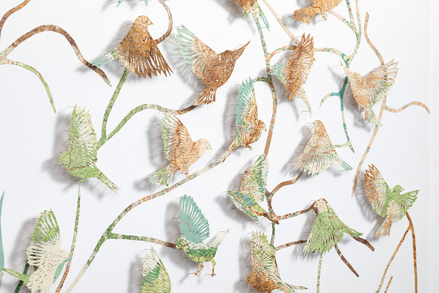 A close up detail of: Seeing The Future by Claire Brewster. A large paper sculpture of birds flying among winding, branch-like paths, hand-cut from pages of a vintage The Times Atlas in earthy green and brown tones.