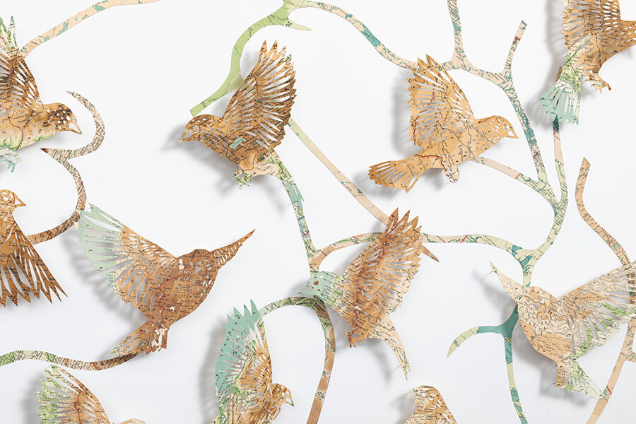 A close up detail of: Seeing The Future by Claire Brewster. A large paper sculpture of birds flying among winding, branch-like paths, hand-cut from pages of a vintage The Times Atlas in earthy green and brown tones.