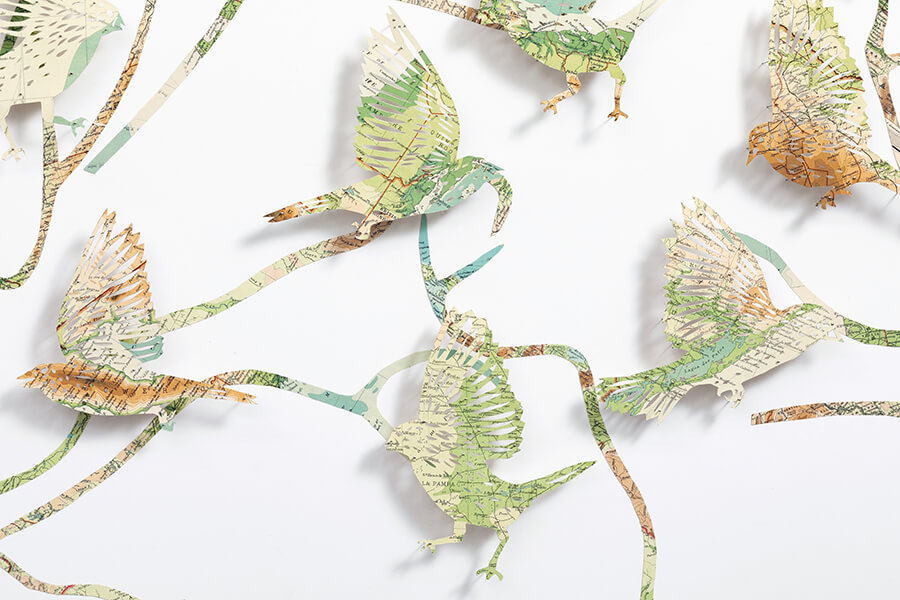 A close up detail of: Seeing The Future by Claire Brewster. A large paper sculpture of birds flying among winding, branch-like paths, hand-cut from pages of a vintage The Times Atlas in earthy green and brown tones.